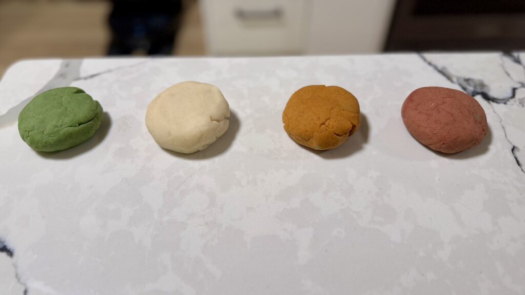 Four balls of scented playdoh lined up on a counter, in varying shades of green, white, orange, and red