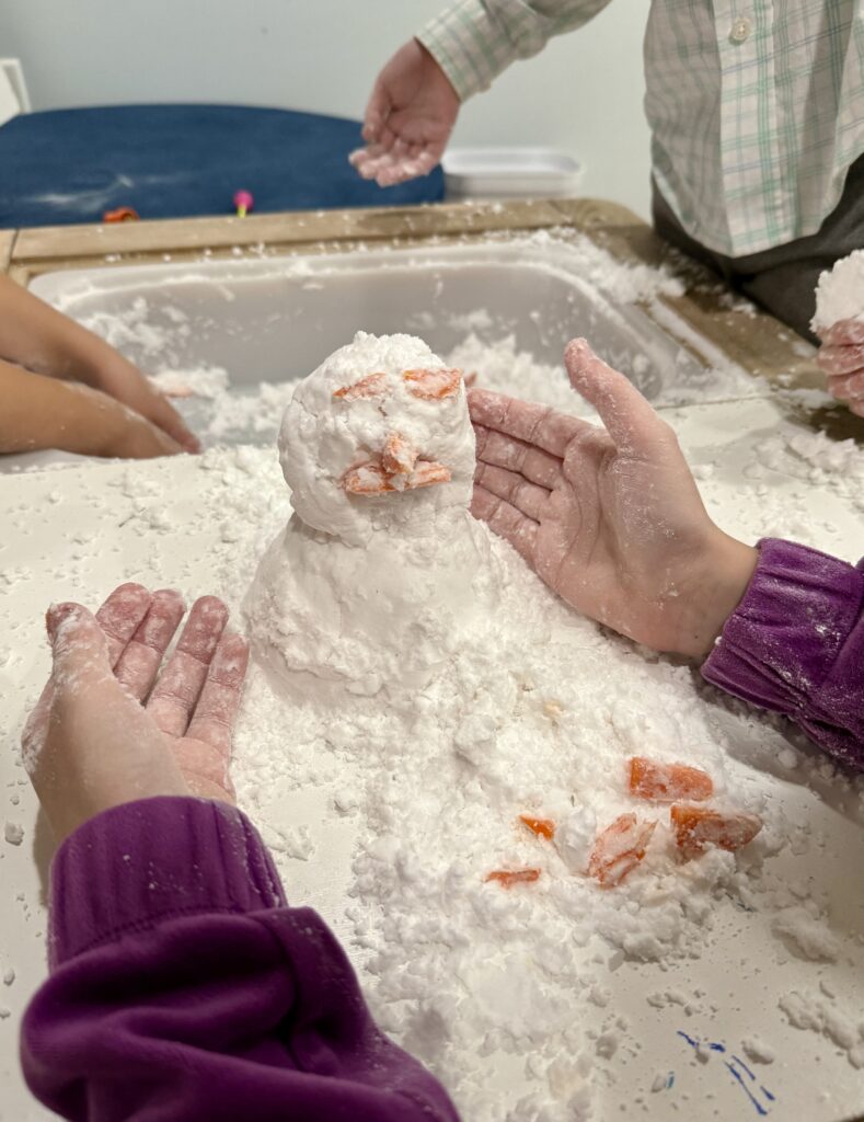 Children's hands around a molded snowman with carrot eyes, nose, and mouth. 