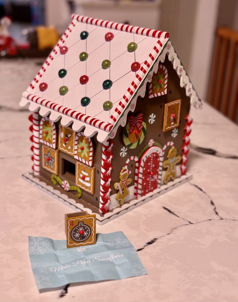 Advent gingerbread house with small wooden boxes for windows and doors, one window pulled out with a paper below that states "Make Paper Snowflakes"