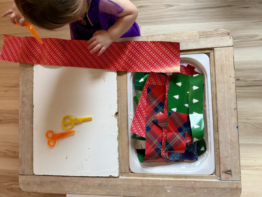 Photo of a child cutting Christmas themed wrapping paper with a handful of scraps in a bin beside her