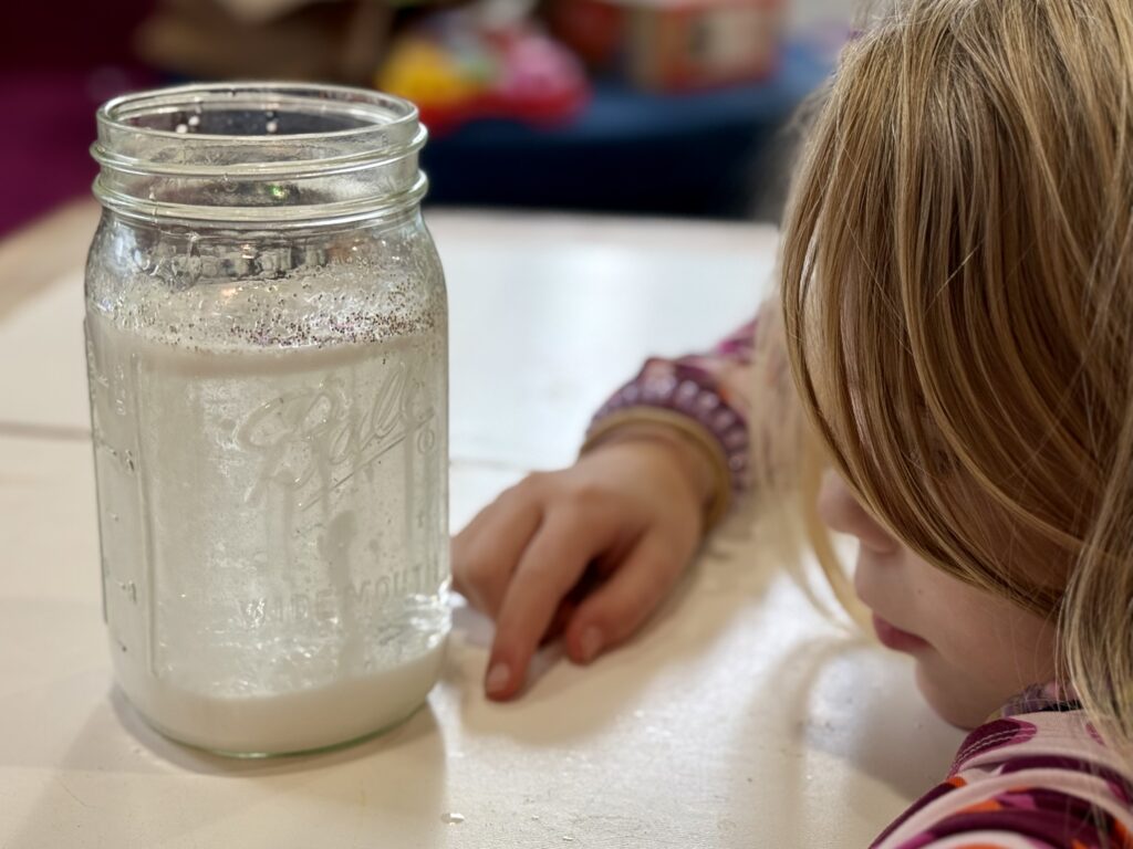 A child looking through a mason jar looking at clear oil and white dyed water creating a snowstorm. 