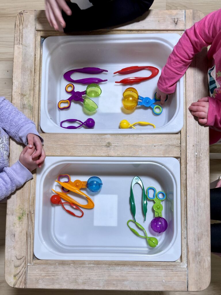 Children's hands in a bin full of colorful fine motor tools: large tweezers, big grabbers, and small squeeze grabs