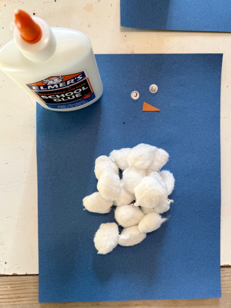 blue construction paper with a bottle of glue, two googly eyes, cotton balls, and an orange triangle on top - the makings for a snowman craft