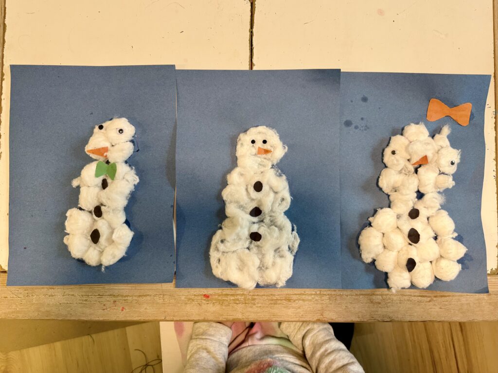 Three pieces of blue construction paper each with a cotton ball snowman on top. Each snowman made from cotton balls with an orange construction paper nose, two googly eyes, and three black buttons, one with a green bow tie and another with an orange bow