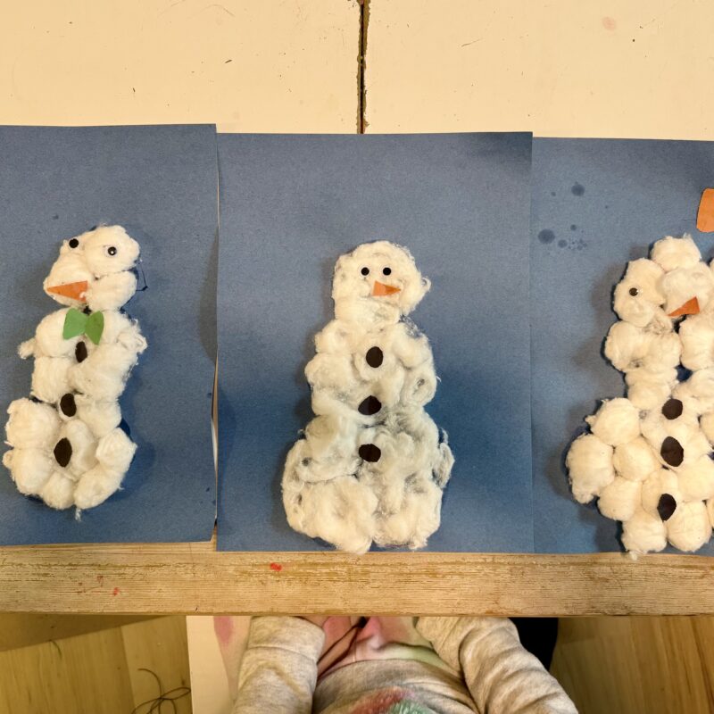 Three pieces of blue construction paper each with a cotton ball snowman on top. Each snowman made from cotton balls with an orange construction paper nose, two googly eyes, and three black buttons, one with a green bow tie and another with an orange bow