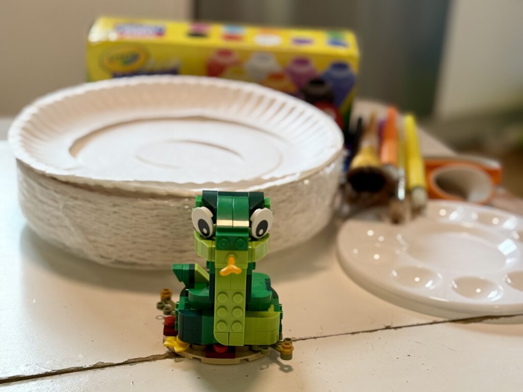 Craft supplies: paper plates, paint, scissors, paintbrushes with a lego snake in the front for the year of the snake