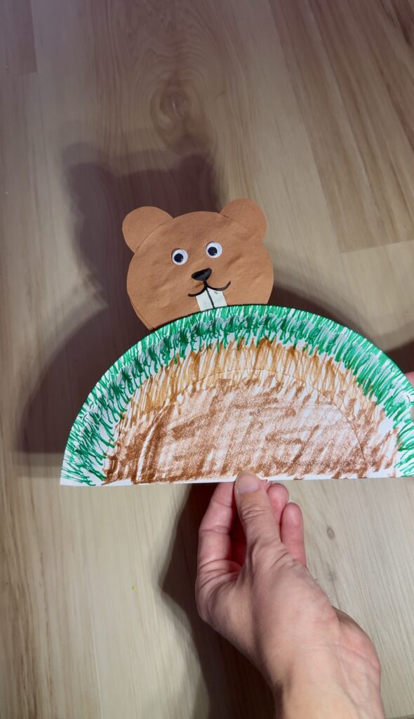 Groundhog Day science experiment: shadow play. Groundhog made of construction paper popping out of a paper plate mound on a popsicle stick. Obvious shadow behind the groundhog.