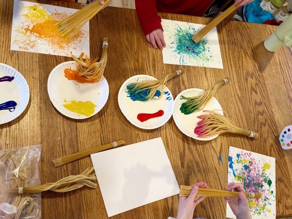 Process art: children painting with paintbrushes made from spaghetti noodles, paper plates with paint on them. 