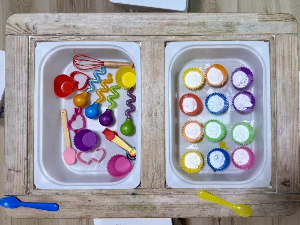 A sensory bin with tools on the left side in a bucket, including: droppers, cookie cutters, whisks, cupcake liners, measuring cups. The other bucket is filled with cupcake liners of all colors filled with baking soda.