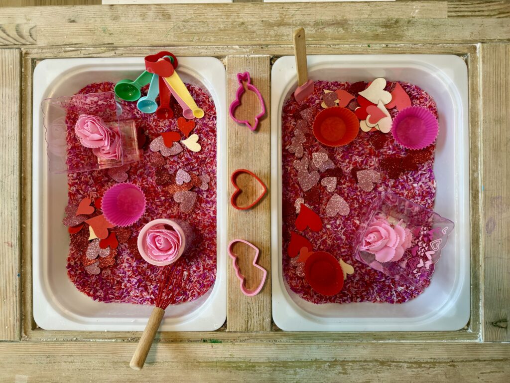 Photo of pink and red rice in a sensory bin filled with heart cookie cutters, pink and red cupcake liners, fake roses, scattered foam and wood hearts, and some utensils to celebrate Valentine's Day. 