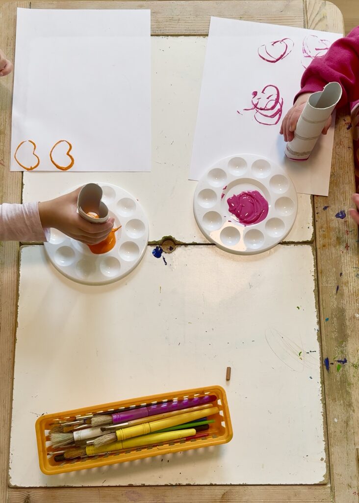 Two kids with paper towel rolls in their hands, shaped like hearts. Placing the heart shape in paint and stamping it onto paper, using the colors orange and pink.