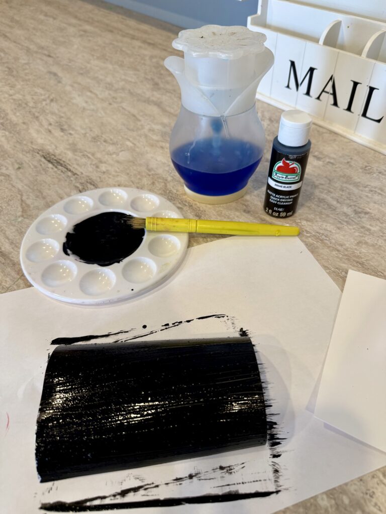 A photo of cardstock painted black with a mix of acrylic paint and dish soap. Yellow paintbrush in a white palette with black paint. Dish soap and black acrylic paint in the background. Creating a Scratch Off Message. 