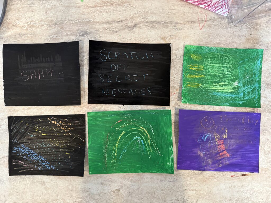 6 pieces of card stock painted black, green, and purple. Hidden messages written in crayons that the paint was scratched to reveal. One black says shh. Another black says scratch off secret messages. Another black rainbow background. A green with a rainbow, a green with popsicles, and a purple with people and message I'm so lucky to be your daughter. 