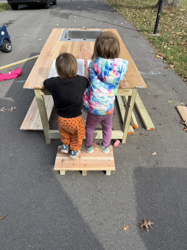 Two kids standing on a stool facing a mud kitchen.