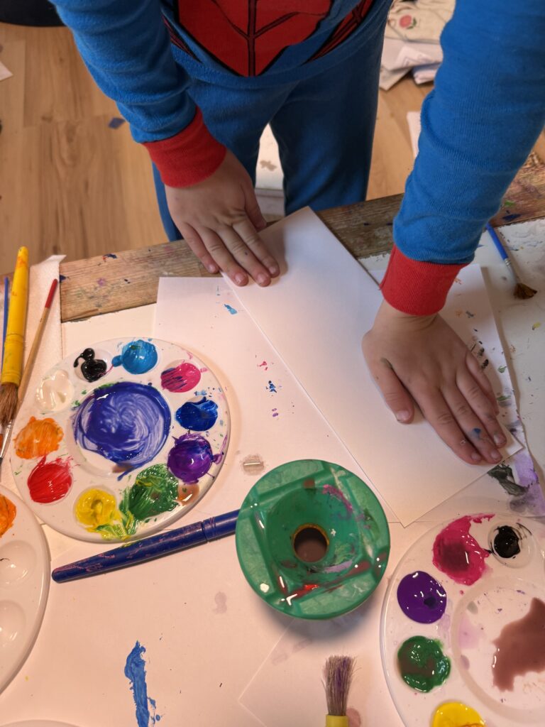 Kids hands pushing down on a folded piece of paper surrounded by painting supplies
