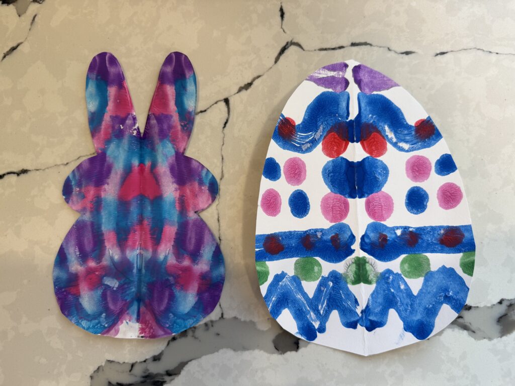 A photo of a bunny and egg with symmetrical patterns of paint made through the process of symmetry art. The bunny is a mixture of pink, blue, and purple. The egg has squiggles, lines, and dots that create patterns in red, blue, pink, and green.