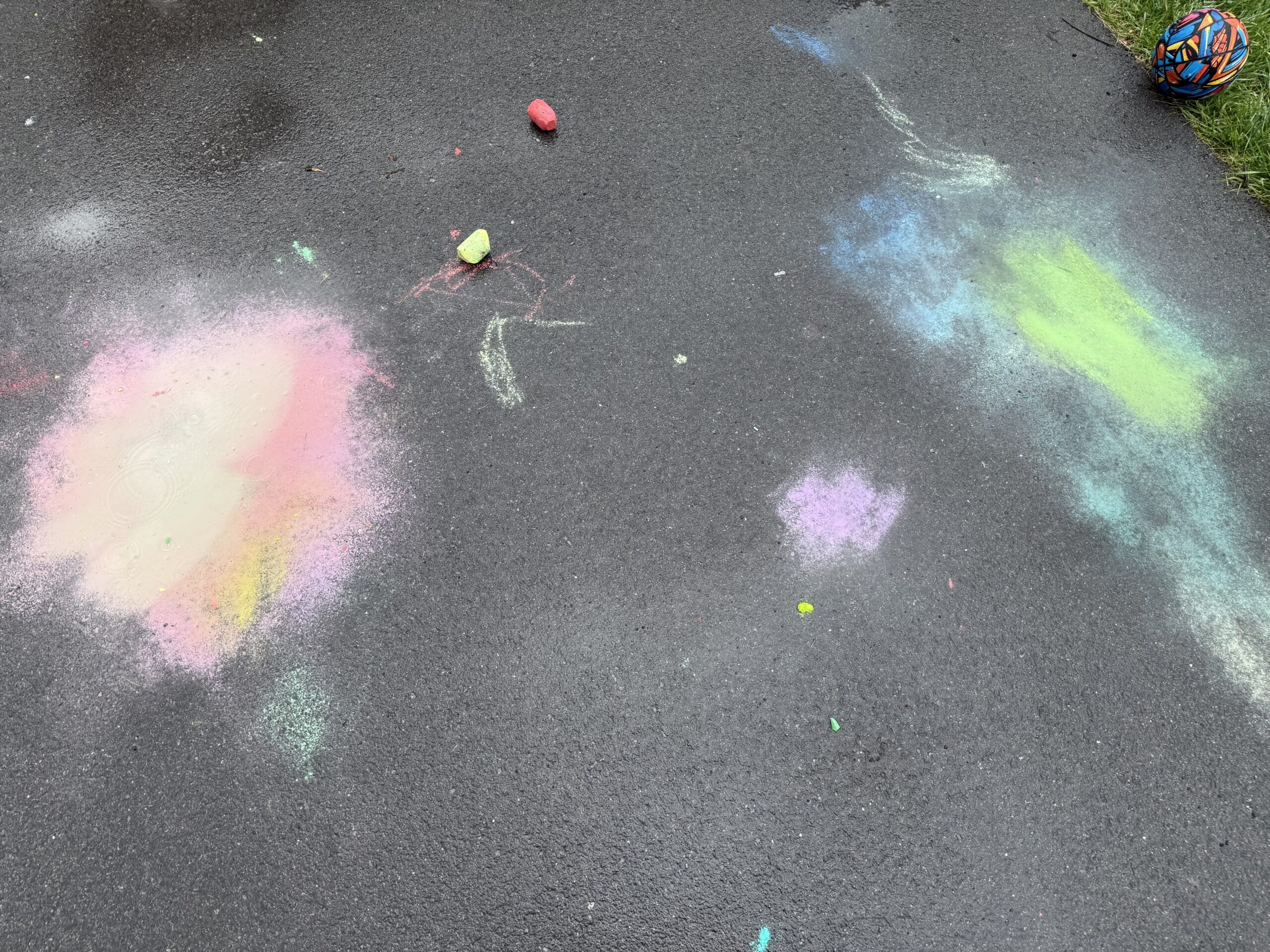 Photo of a driveway with some chalk filled puddles. One on the left in pink and yellow, one on the right in green and blue.