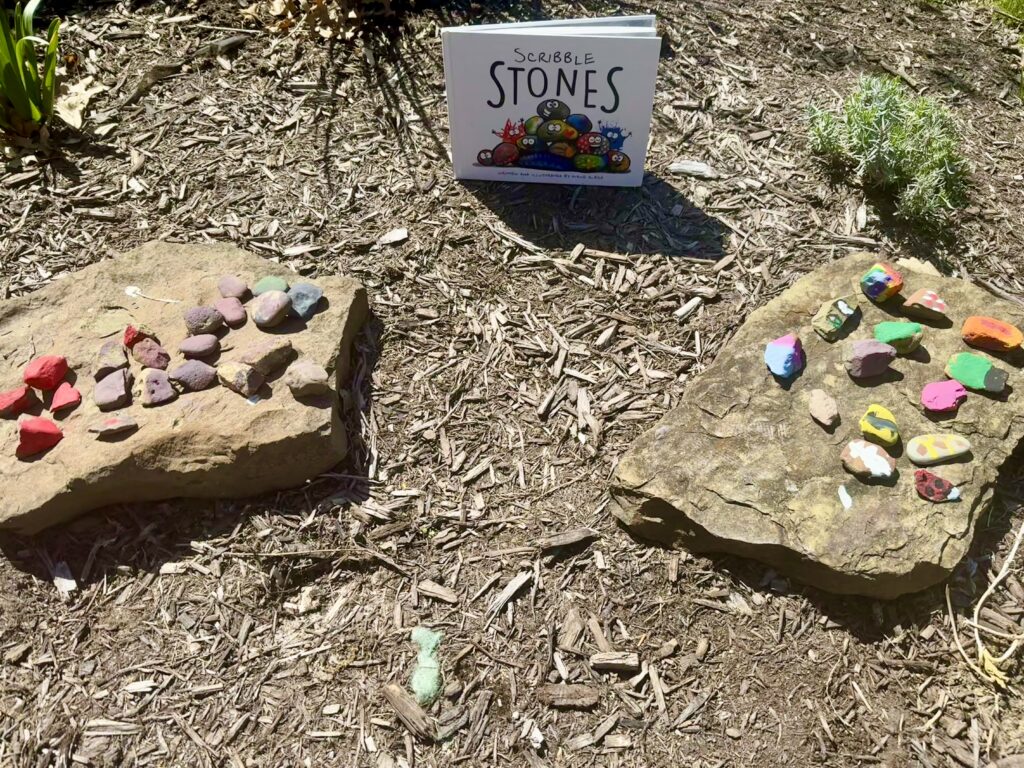 Painted rocks sitting on larger rocks beside the book "Scribble Stones" by Diane Alber.